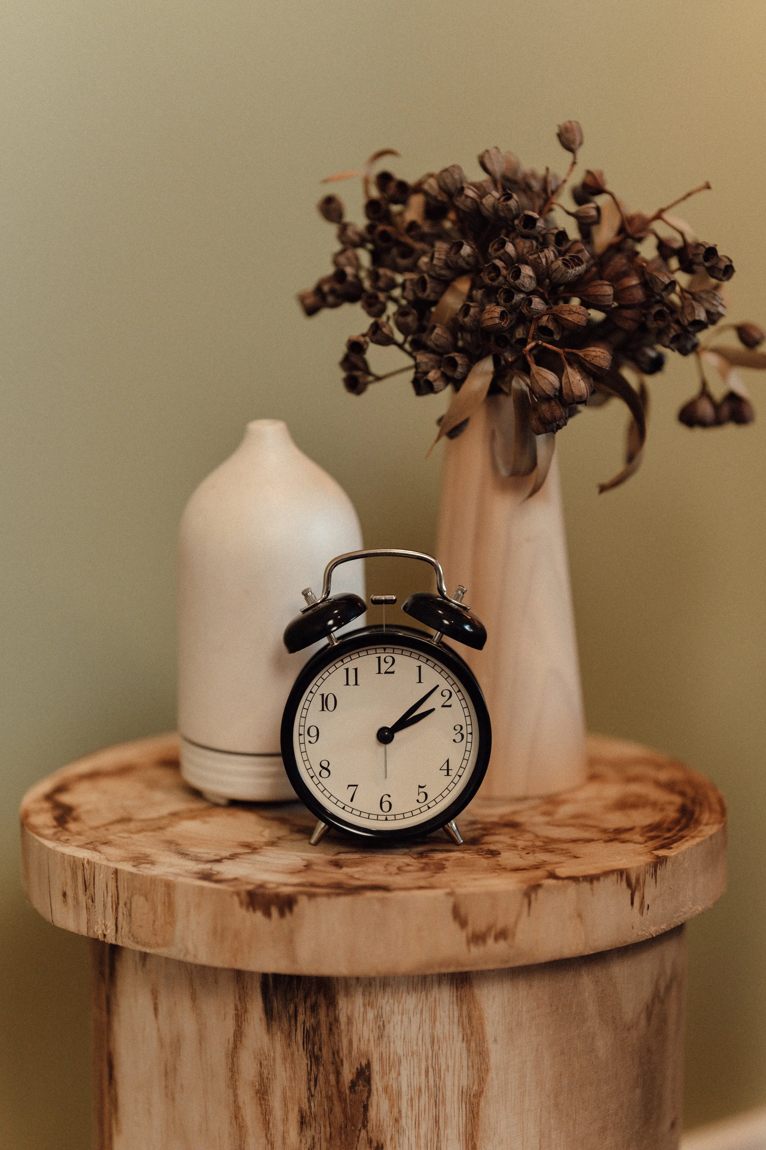 A round wooden table with a small black alarm clock showing 1:08, a white ceramic vase, and a taller beige ceramic vase holding dried flowers against a light green wall.