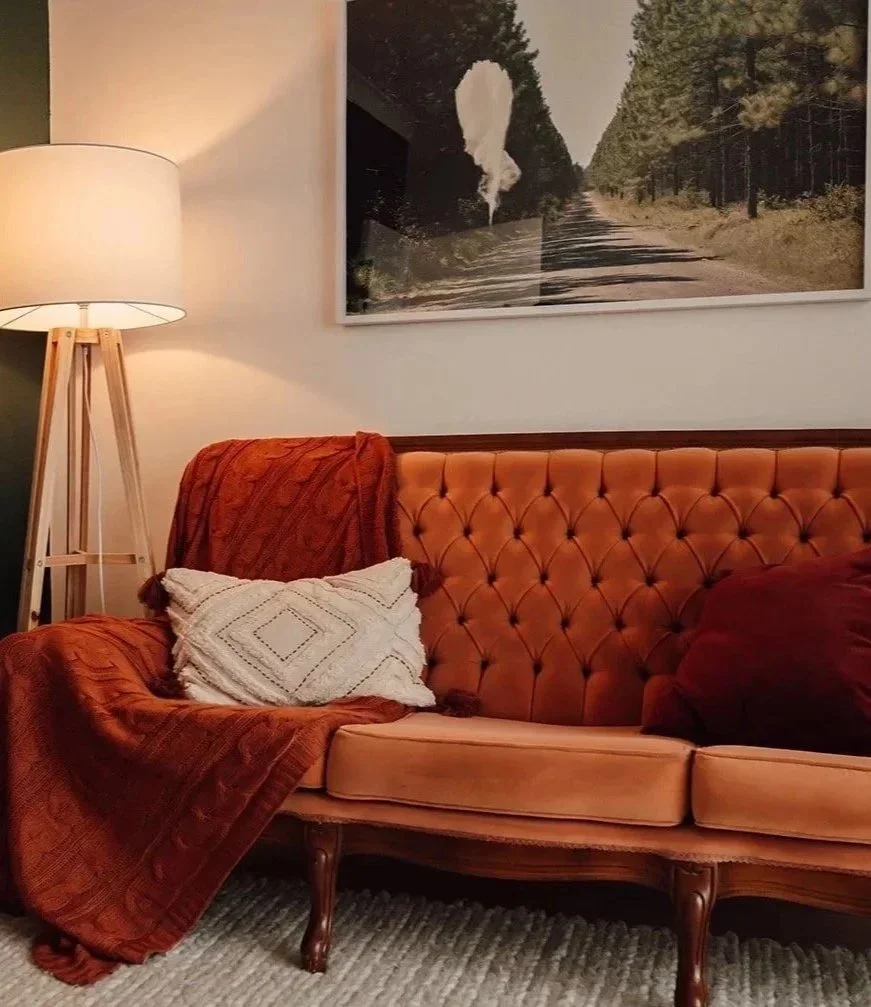 A vintage orange tufted sofa with pillows and a folded blanket, illuminated by a tall floor lamp, in a cozy therapy room