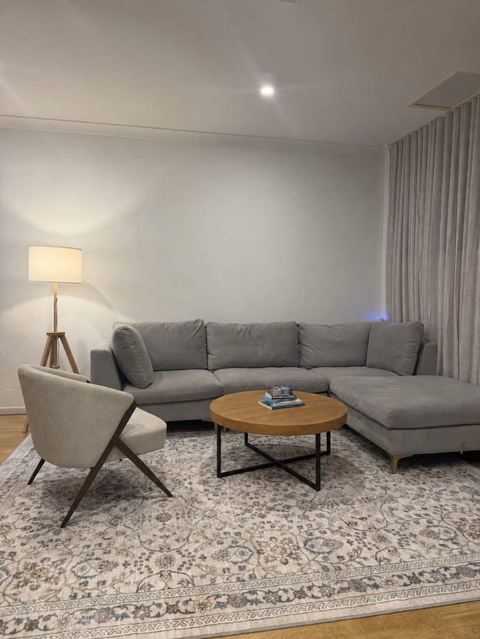 Consulting room with a large grey corner lounge, a pale armchair, a round timber coffee table and a patterned rug beside a tripod floor lamp