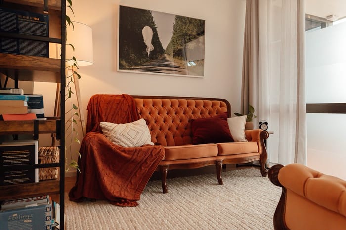 Consulting room with a burnt orange velvet lounge, a knitted rust throw and cushions, a timber shelf of books and baskets, a floor lamp and a framed landscape print